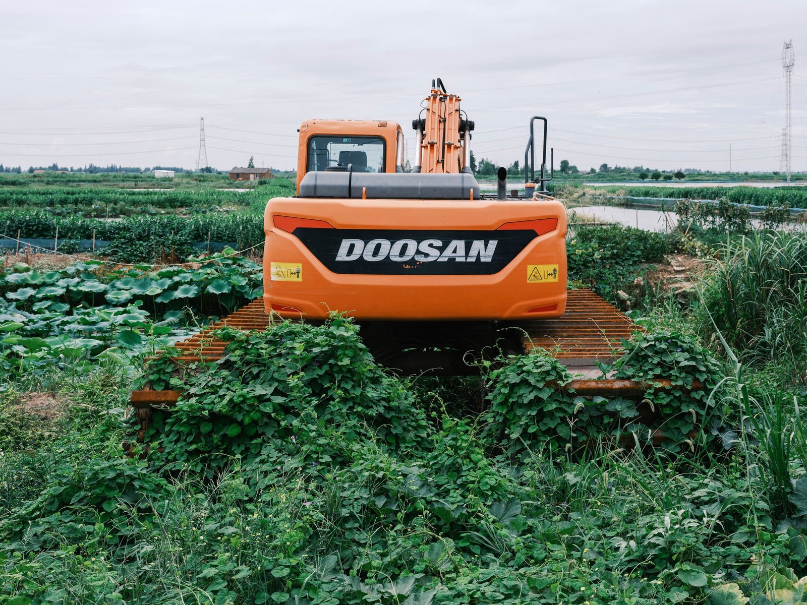 Home Bright orange excavator working in a lush green agricultural field under a cloudy sky.